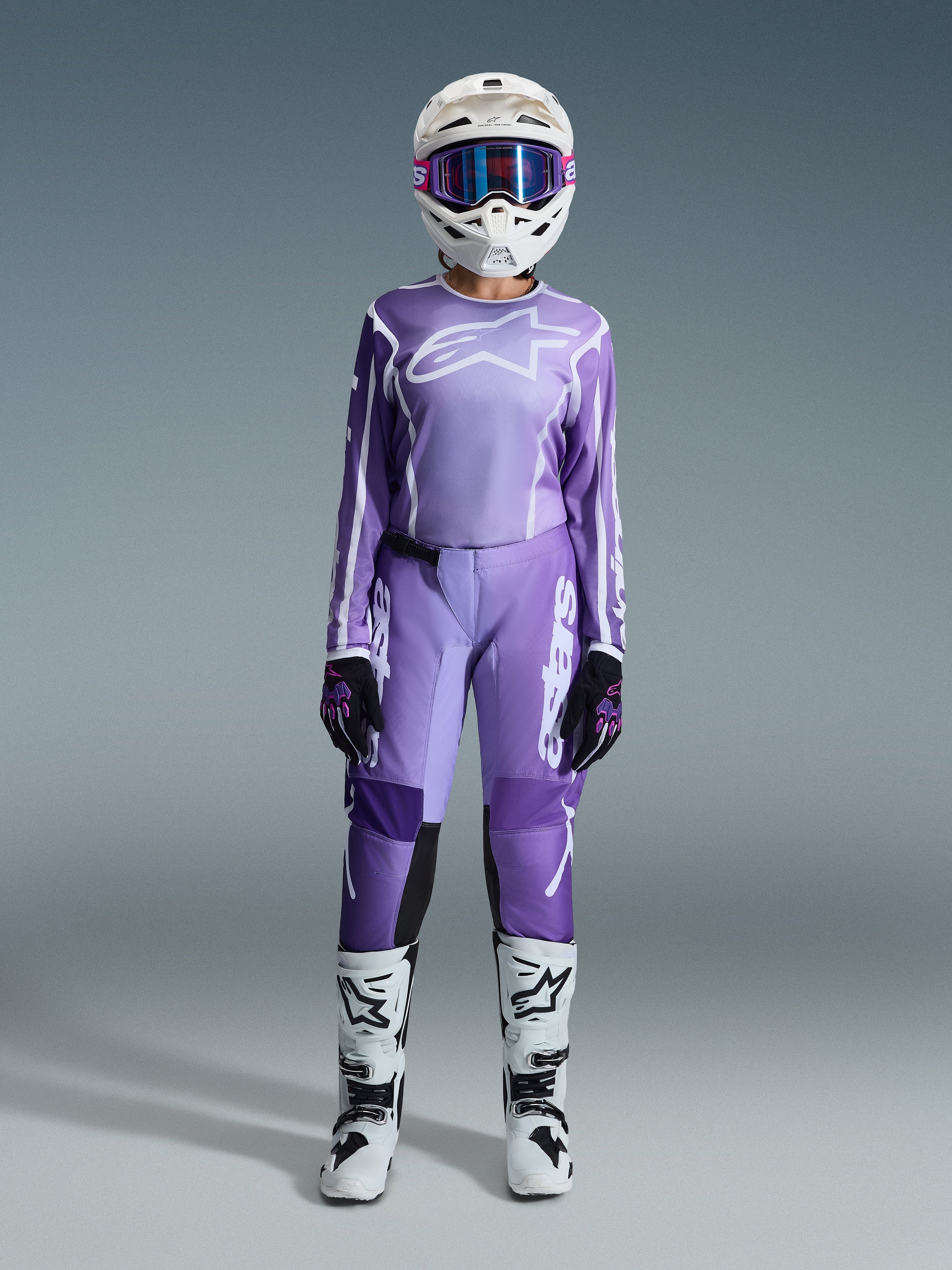 Piloto de motocross com o Conjunto Alpinestars Stella Fluid Apex Feminino 2026, camisola violeta e branca com calças a condizer, capacete branco com óculos cor-de-rosa e botas de todo-o-terreno brancas e pretas, kit de corrida completo para motocross feminino