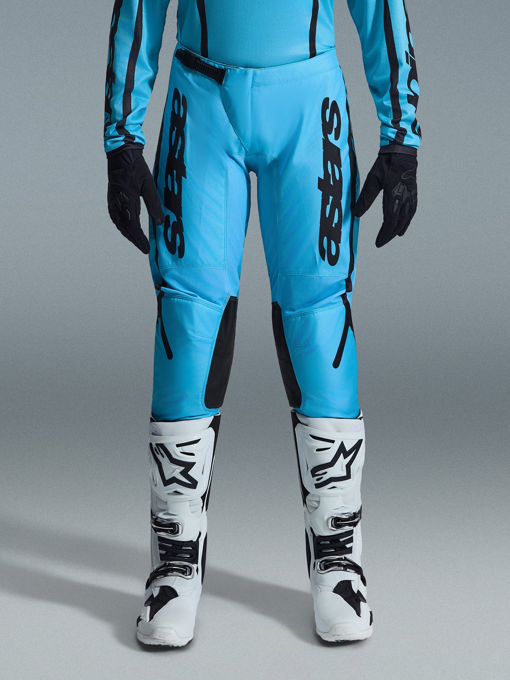 Calças Alpinestars Stella Fluid Apex Femininas 2026, calças de motocross, azul ciano e preto, logótipo preto vertical nas coxas, painéis reforçados na parte interna dos joelhos em preto, apresentadas com botas de motocross brancas e pretas e luvas pretas, equipamento profissional de todo-o-terreno para mulher