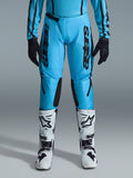 Calças Alpinestars Stella Fluid Apex Femininas 2026, calças de motocross, azul ciano e preto, logótipo preto vertical nas coxas, painéis reforçados na parte interna dos joelhos em preto, apresentadas com botas de motocross brancas e pretas e luvas pretas, equipamento profissional de todo-o-terreno para mulher