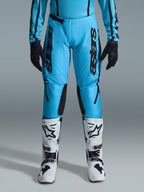 Calças Alpinestars Stella Fluid Apex Femininas 2026, calças de motocross, azul ciano e preto, logótipo preto vertical nas coxas, painéis reforçados na parte interna dos joelhos em preto, apresentadas com botas de motocross brancas e pretas e luvas pretas, equipamento profissional de todo-o-terreno para mulher
