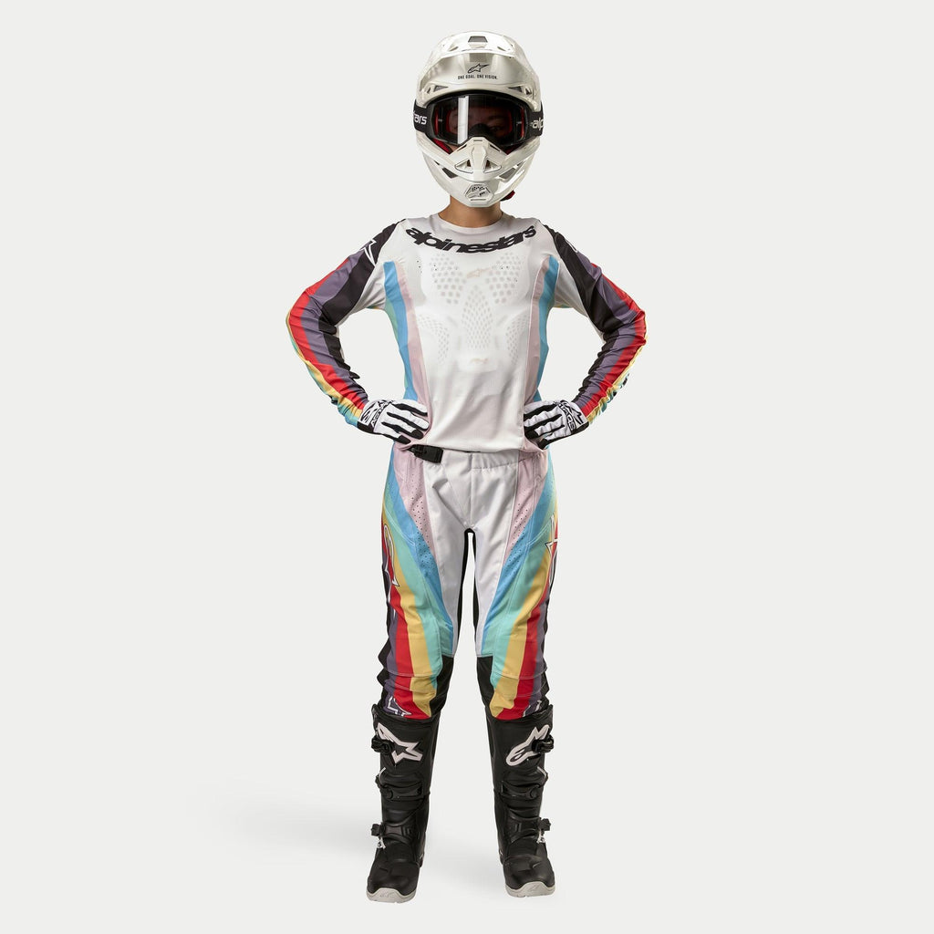 Piloto de motocross feminina a usar calças Alpinestars Stella Techstar 2024, Black Multicolor com base branca e riscas vermelhas, amarelas e azuis, camisola de manga comprida a condizer, capacete branco e botas pretas, equipamento de corrida completo para competição todo-o-terreno