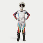 Piloto de motocross feminina a usar calças Alpinestars Stella Techstar 2024, Black Multicolor com base branca e riscas vermelhas, amarelas e azuis, camisola de manga comprida a condizer, capacete branco e botas pretas, equipamento de corrida completo para competição todo-o-terreno