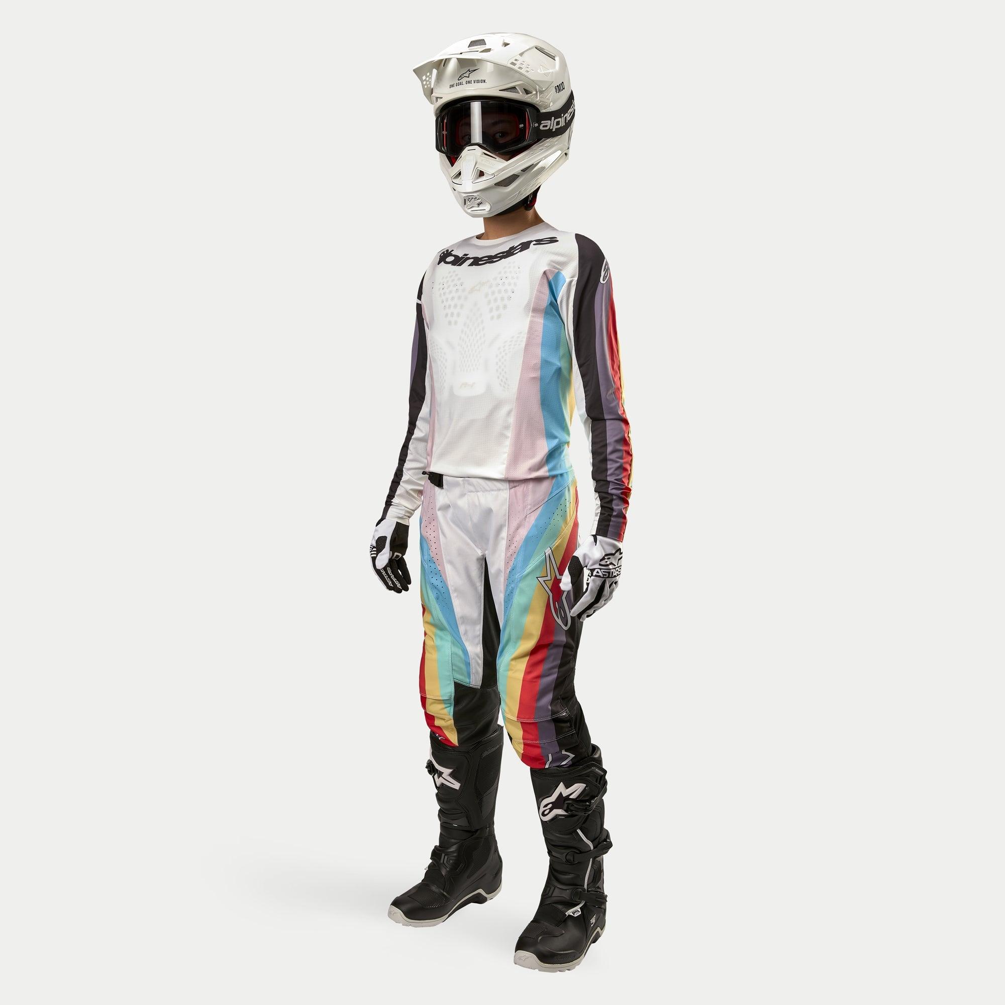 Piloto de motocross a usar calças Alpinestars Stella Techstar Femininas 2024, calças de motocross, preto multicolor com painéis brancos e riscas diagonais vibrantes, camisola a condizer, capacete branco e botas pretas, conjunto de corrida todo-o-terreno coordenado para mulher