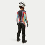 Piloto de motocross vista de costas a usar calças Alpinestars Stella Techstar Femininas 2024 em Preto Multicor, com logótipo branco de grandes dimensões na zona posterior, combinadas com camisola às riscas coloridas, capacete branco e botas Tech 3 pretas para todo-o-terreno