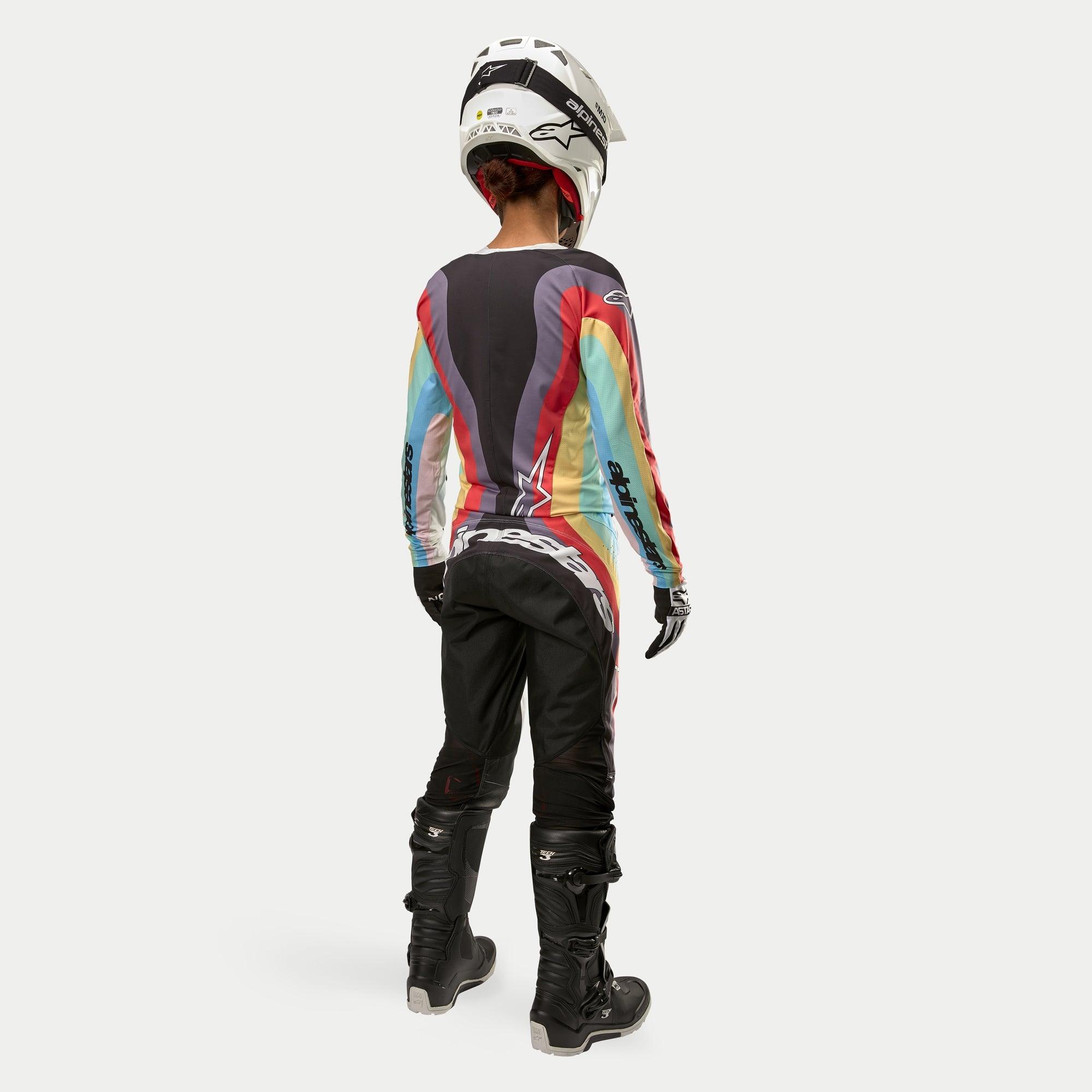 Piloto de motocross vista de costas a usar calças Alpinestars Stella Techstar Femininas 2024 em Preto Multicor, com logótipo branco de grandes dimensões na zona posterior, combinadas com camisola às riscas coloridas, capacete branco e botas Tech 3 pretas para todo-o-terreno