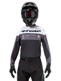 Piloto de motocross a usar a camisola Alpinestars Pro-Dura em Black Magnet White, camisola de performance de manga comprida com blocos de cor brancos e cinzentos, usada com um capacete e óculos pretos, com a marca 'alpinestars' e o slogan 'ONE GOAL ONE VISION', vestuário de corrida de motociclismo todo-o-terreno