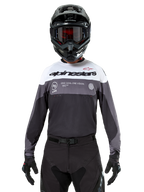 Piloto de motocross a usar a camisola Alpinestars Pro-Dura em Black Magnet White, camisola de performance de manga comprida com blocos de cor brancos e cinzentos, usada com um capacete e óculos pretos, com a marca 'alpinestars' e o slogan 'ONE GOAL ONE VISION', vestuário de corrida de motociclismo todo-o-terreno