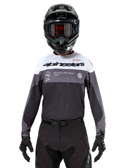 Motocross rider wearing the Alpinestars Pro-Dura Jersey in Black Magnet White, long-sleeve performance top with white and grey color blocks, worn with a black helmet and goggles, featuring 'alpinestars' branding and 'ONE GOAL ONE VISION' slogan, off-road motorcycle racing apparel