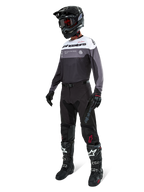 Camisola Alpinestars Pro-Dura em Black Magnet White, vestida por um piloto de motocross com equipamento completo, incluindo calças pretas, capacete preto com óculos e botas pretas e brancas. A camisola apresenta um design técnico de blocos de cor com painéis brancos, cinzentos e pretos, concebida para performance em enduro e todo-o-terreno.