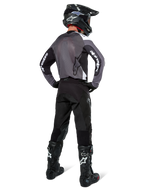 Piloto de motocross com camisola Alpinestars Pro-Dura em Black Magnet White, vista traseira mostrando painel traseiro em malha cinzenta e mangas pretas com marca Alpinestars branca, coordenada com calças pretas, capacete preto e botas pretas e brancas, equipamento profissional de enduro
