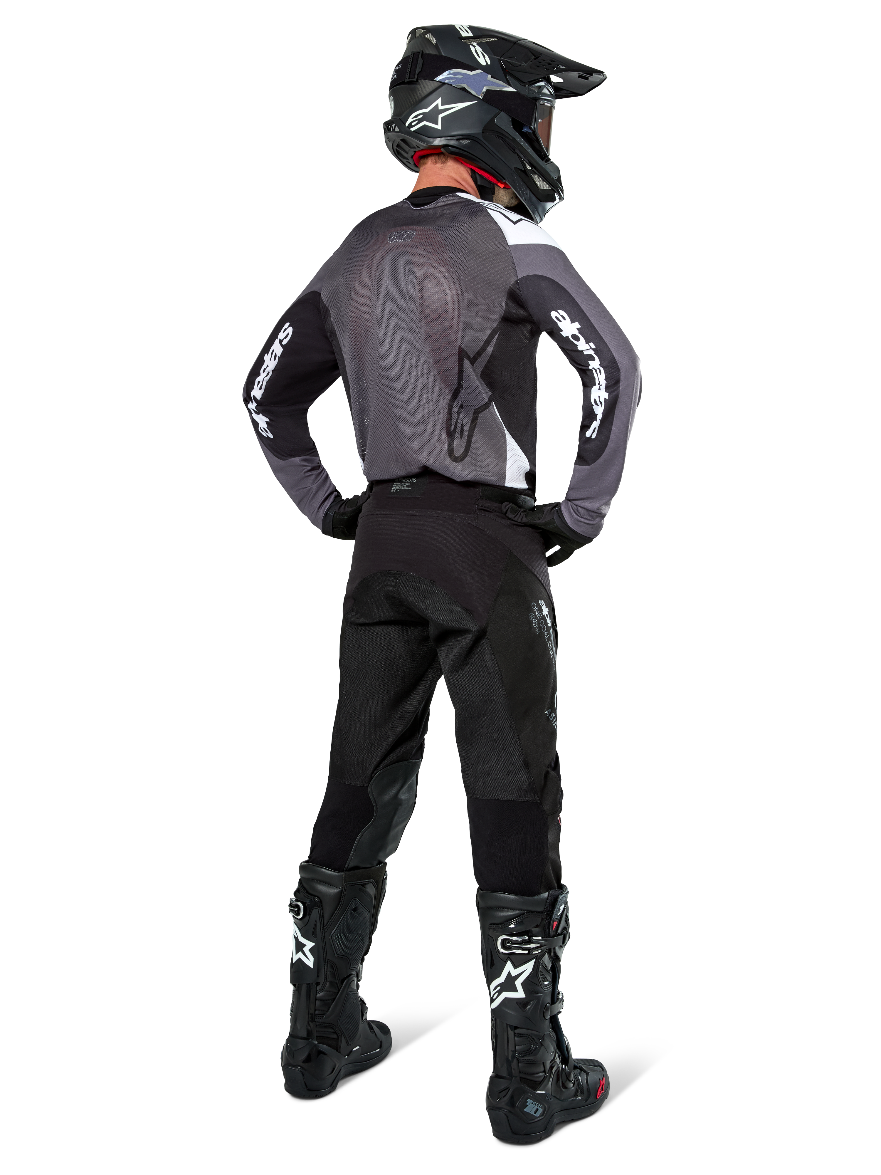 Piloto de motocross com camisola Alpinestars Pro-Dura em Black Magnet White, vista traseira mostrando painel traseiro em malha cinzenta e mangas pretas com marca Alpinestars branca, coordenada com calças pretas, capacete preto e botas pretas e brancas, equipamento profissional de enduro