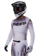 Motocross rider wearing the Alpinestars 2025 Supertech Maker Gear Set in White, featuring a grey gradient jersey with the black astars logo and laser-cut perforations, matching pants with magnetic buckle and star graphics, worn with a black and white helmet and gloves for a complete off-road racing look.