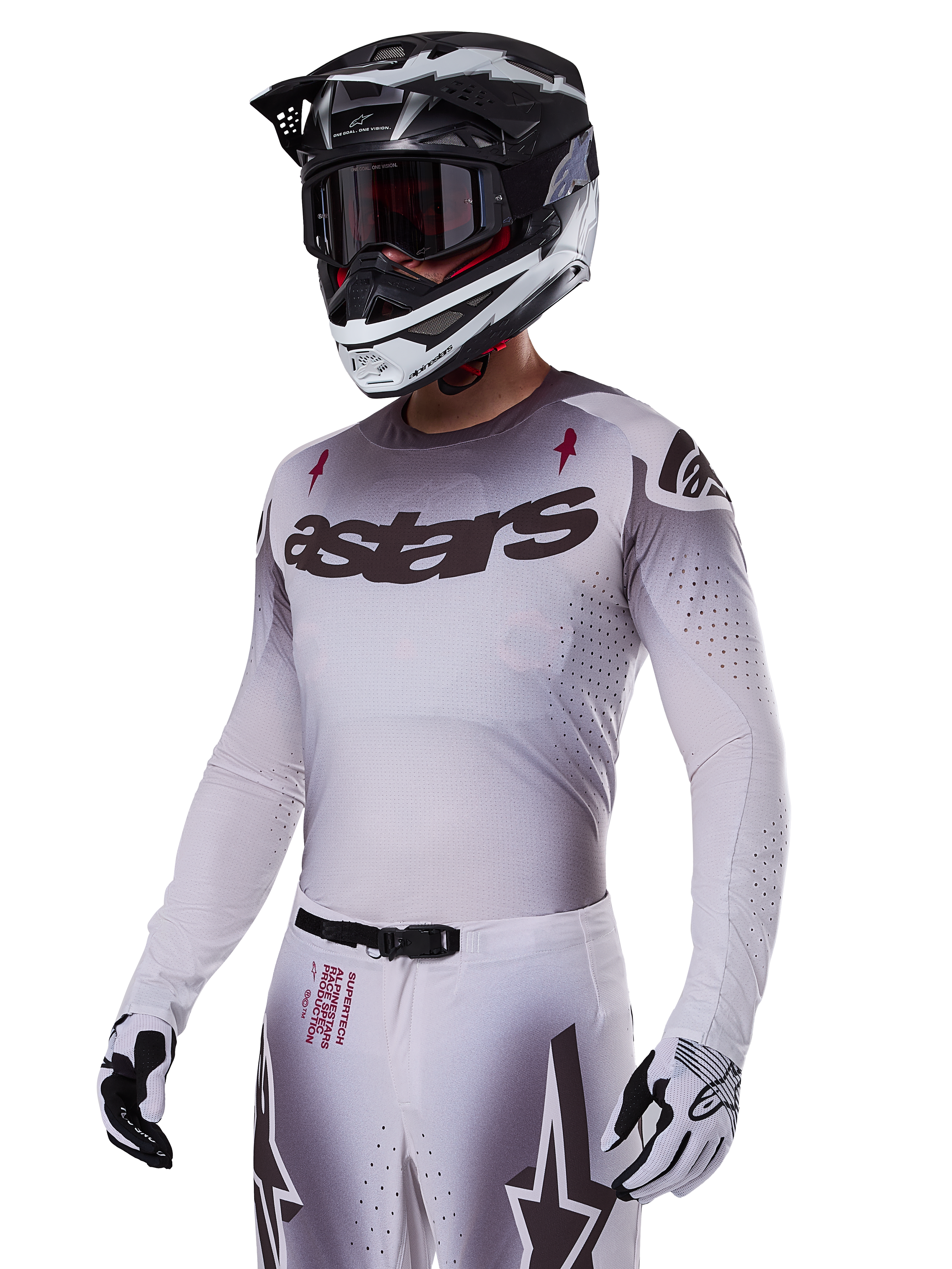 Motocross rider wearing the Alpinestars 2025 Supertech Maker Gear Set in White, featuring a grey gradient jersey with the black astars logo and laser-cut perforations, matching pants with magnetic buckle and star graphics, worn with a black and white helmet and gloves for a complete off-road racing look.
