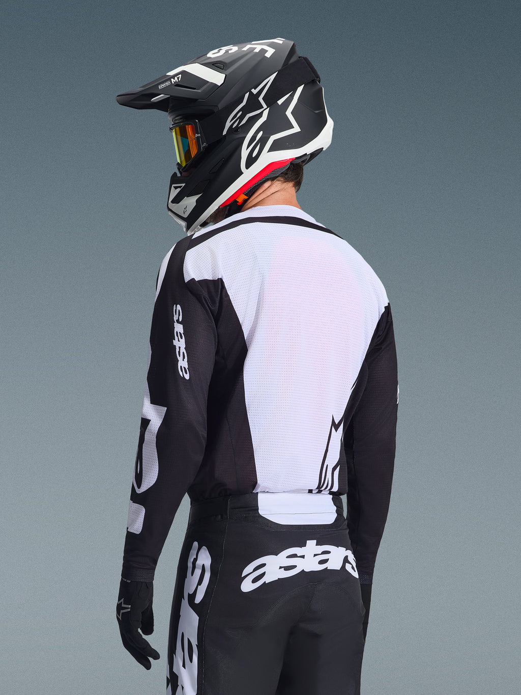 Piloto de motocross visto de costas a usar a camisola Alpinestars 2026 Racer Air Riway, em preto e branco, com um painel traseiro em malha respirável branca e mangas pretas com a marca 'astars', calças pretas a condizer com logótipos brancos, capacete preto com detalhes em branco, equipamento profissional de todo-o-terreno para desempenho em temperaturas elevadas