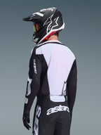 Piloto de motocross visto de costas a usar a camisola Alpinestars 2026 Racer Air Riway, em preto e branco, com um painel traseiro em malha respirável branca e mangas pretas com a marca 'astars', calças pretas a condizer com logótipos brancos, capacete preto com detalhes em branco, equipamento profissional de todo-o-terreno para desempenho em temperaturas elevadas