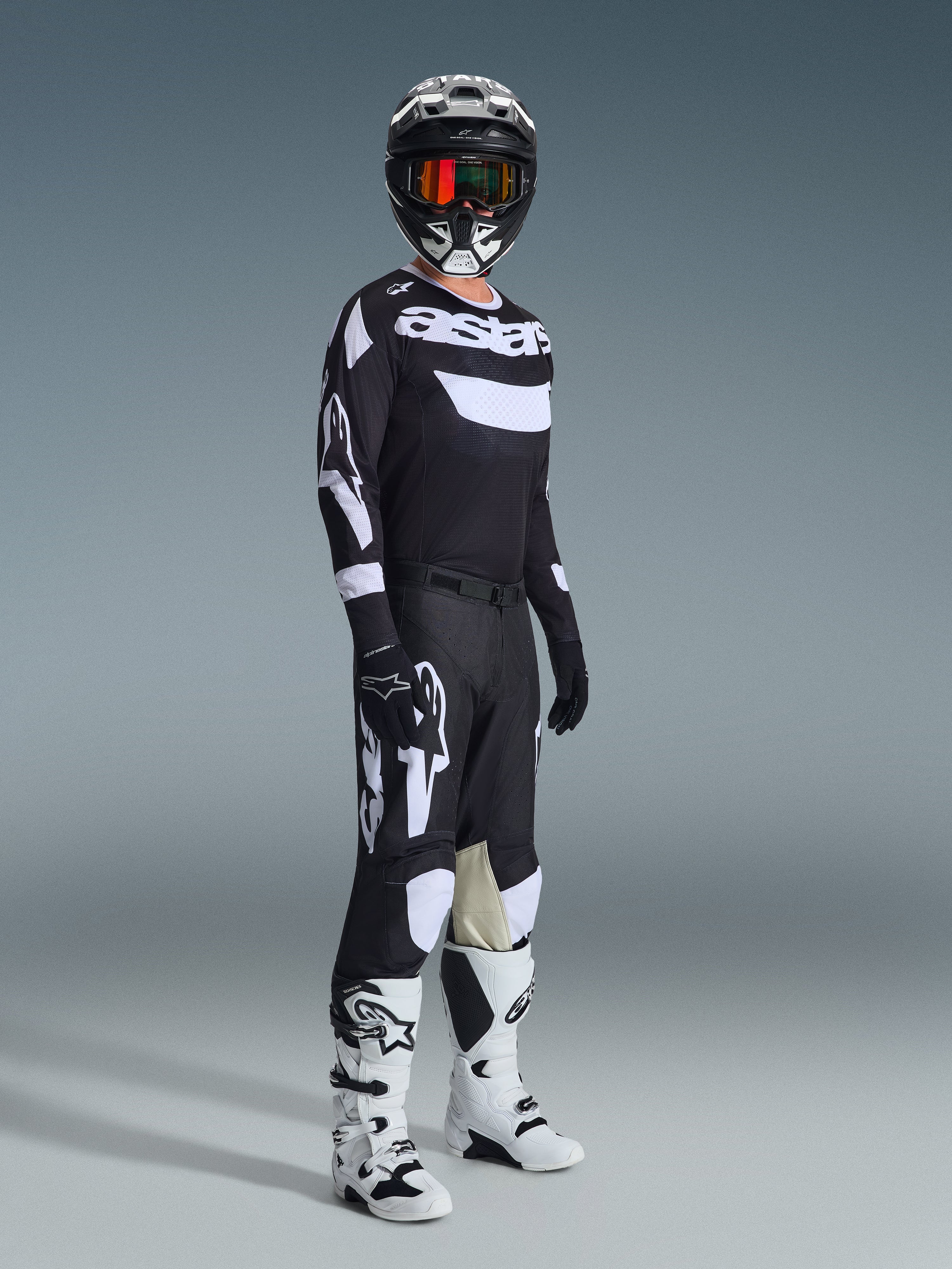 Piloto de motocross com equipamento de corrida completo, Camisola Racer Air Riway 2026 em preto e branco, calças pretas com grandes logótipos Alpinestars brancos, capacete preto com detalhes brancos e óculos cor de laranja, botas brancas e pretas, conjunto coordenado para corridas profissionais de todo-o-terreno