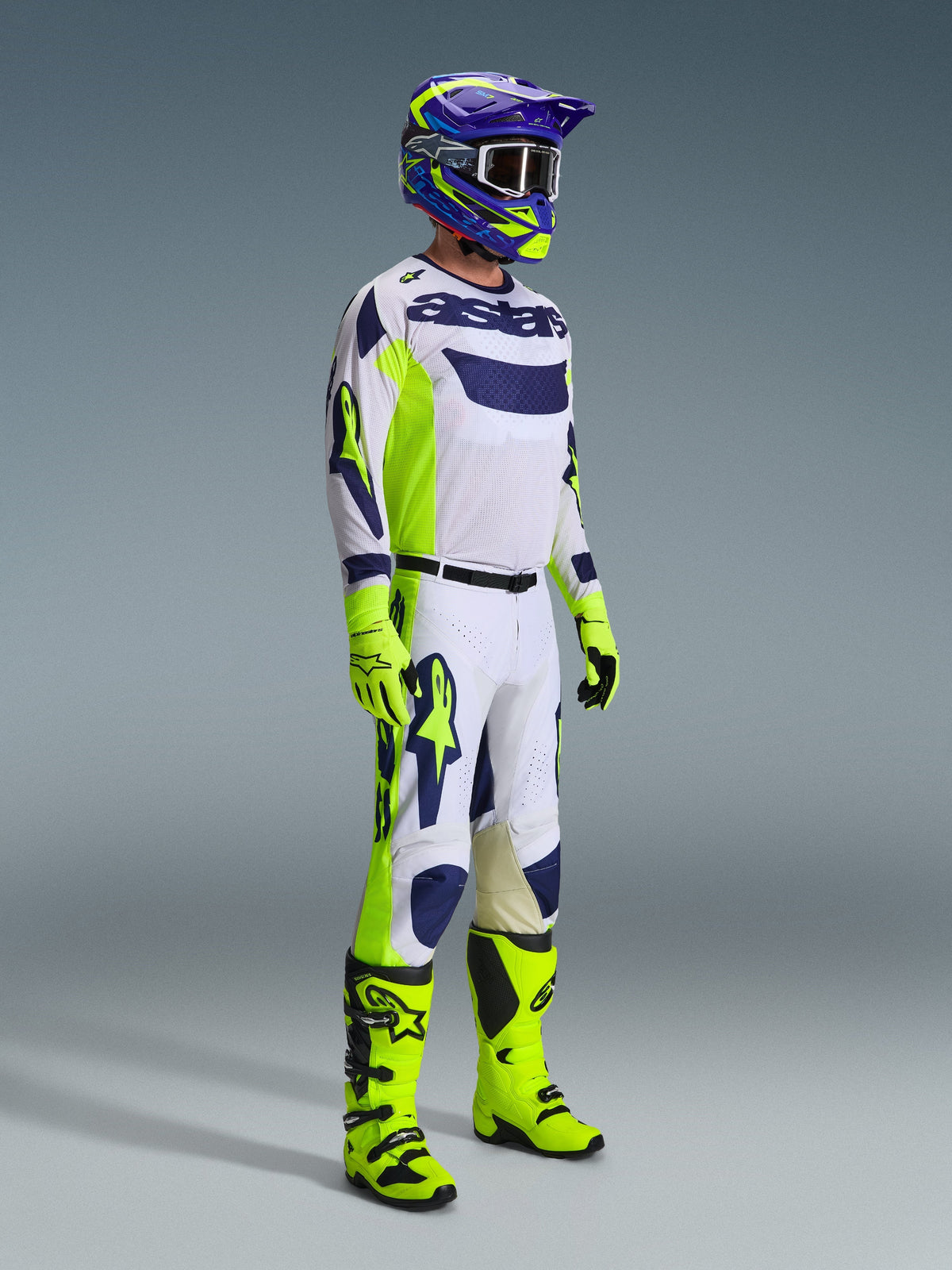 Piloto de motocross a usar o Conjunto de Equipamento Alpinestars 2026 Racer Air Riway em Cinzento Claro, Amarelo Fluo e Azul. O conjunto inclui uma camisola de malha de manga comprida, calças de corrida a condizer, luvas e botas em amarelo néon vibrante, e um capacete roxo e azul com óculos, concebido para um desempenho off-road ventilado.