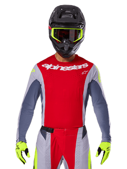 Motocross rider wearing 2025 Techstar Melt Jersey and matching pants, Bright Red and Gray with white Alpinestars branding, black helmet with neon yellow goggles, matching neon yellow gloves, ventilated performance gear for off-road racing