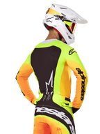 Piloto de motocross com camisola Alpinestars 2025 Techstar Melt, vista traseira, amarelo néon com mangas cor de laranja e um grande painel gráfico preto, calças de motocross a condizer com logótipo proeminente, capacete Alpinestars branco e óculos cor de laranja, equipamento profissional de corrida todo-o-terreno