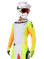 Piloto de motocross a usar o conjunto Alpinestars 2025 Techstar Melt, camisola branca com mangas em amarelo vibrante e laranja, logótipo 'Alpinestars' preto no peito, luvas em amarelo néon, capacete branco com óculos de lentes vermelhas, kit profissional de corrida para todo-o-terreno