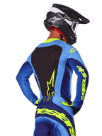 Vista traseira de um piloto de motocross a usar a Camisola Alpinestars 2025 Techstar Melt em azul e amarelo fluo, com painel traseiro preto e logótipo amarelo destacado nas mangas, combinada com calças de motocross a condizer e um capacete de carbono preto, equipamento de performance para corridas off-road