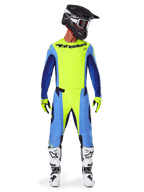 Piloto de motocross com equipamento de corrida completo, camisola Alpinestars 2025 Techstar Melt em Amarelo Fluo e Azul, calças de motocross a condizer em amarelo e azul, capacete preto com grafismos brancos e óculos, botas brancas e pretas, luvas amarelas e pretas, conjunto de performance para todo-o-terreno