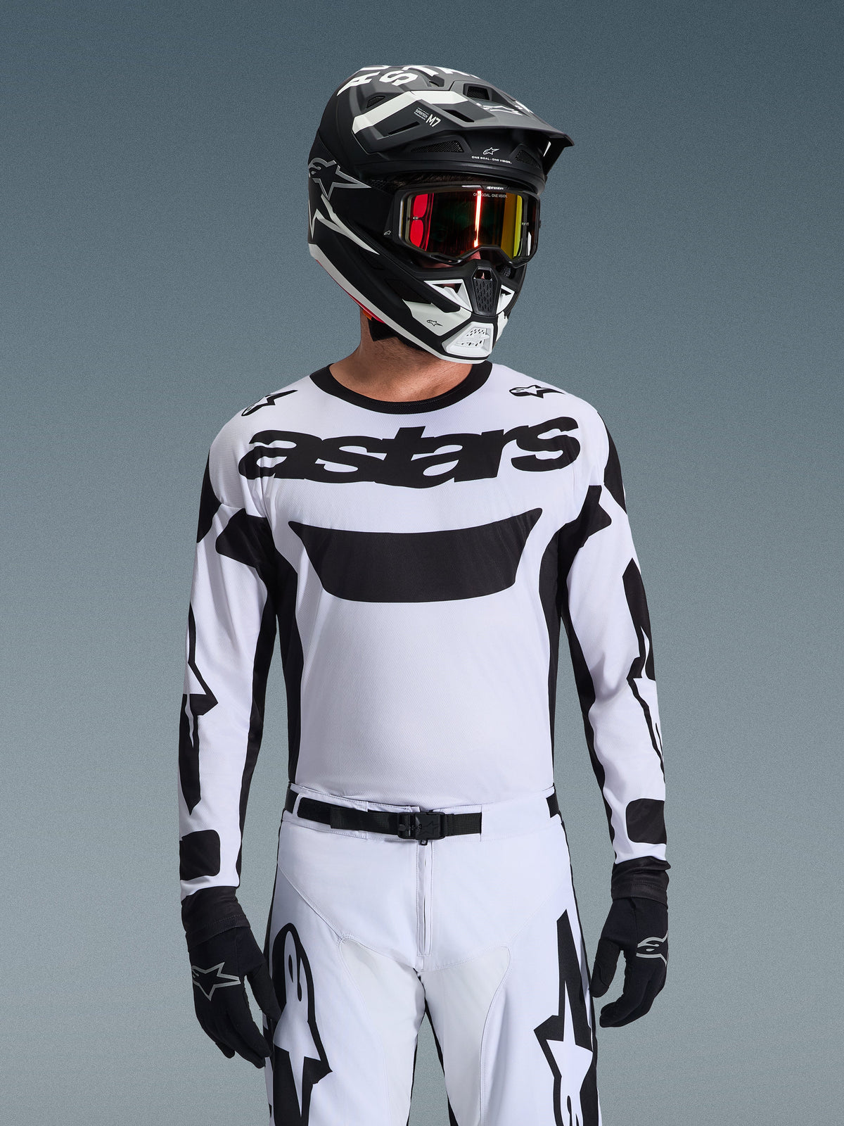 Piloto de motocross a usar a camisola Alpinestars 2026 Racer Riway em branco e preto, com a marca 'astars' no peito, calças brancas e pretas a condizer, capacete preto com óculos e luvas pretas, equipamento coordenado para corridas todo-o-terreno
