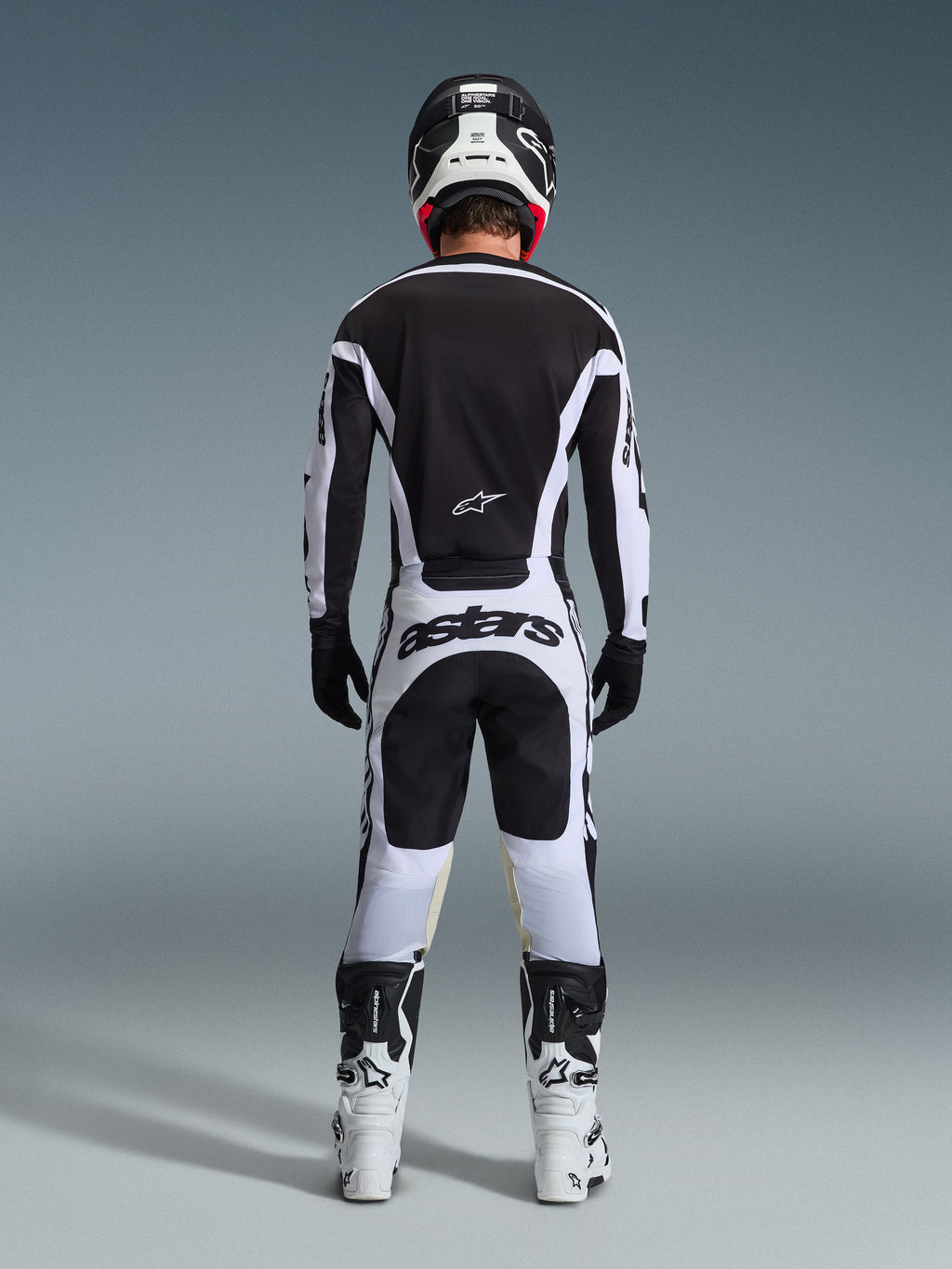 Vista traseira de um piloto de motocross a usar o Conjunto de Equipamento Racer Riway 2026 em preto e branco, composto por camisola preta com detalhes brancos, calças brancas com a marca 'astars' no assento, capacete preto, botas brancas e luvas pretas, equipamento profissional completo de corrida todo-o-terreno