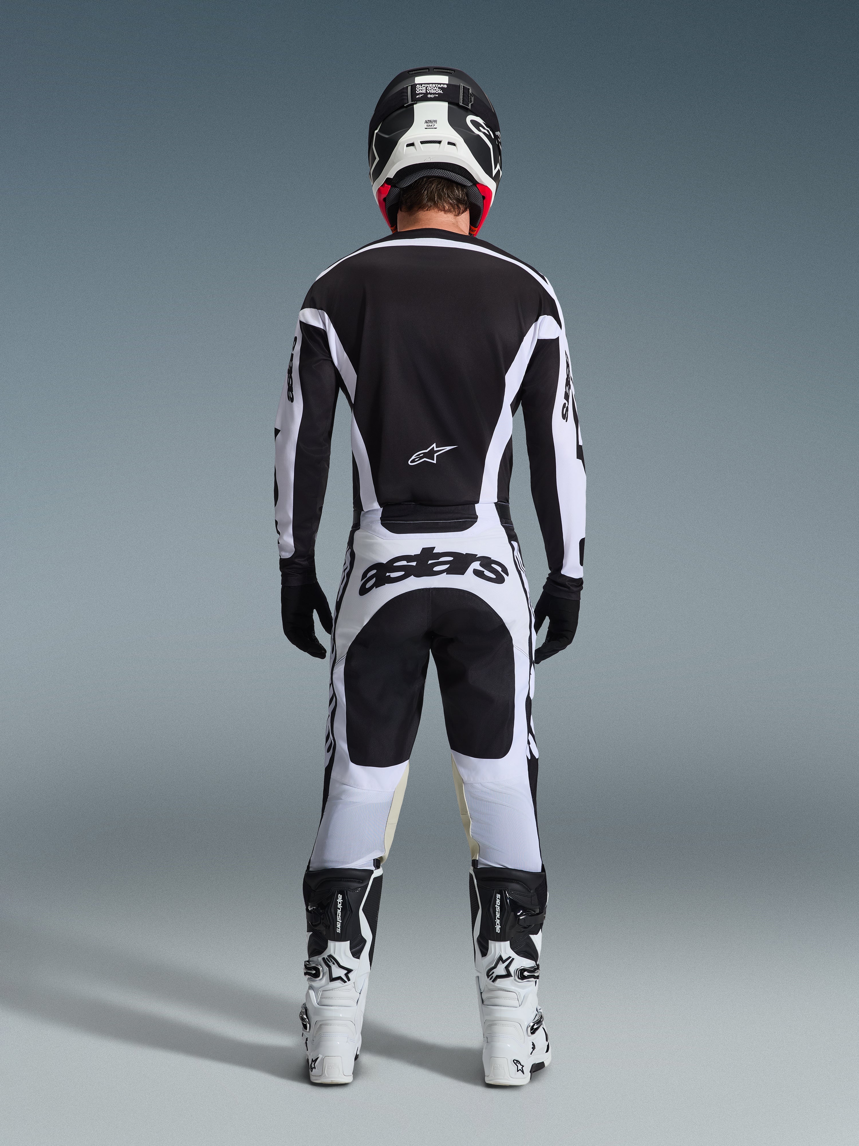 Vista traseira de um piloto de motocross a usar o Conjunto de Equipamento Racer Riway 2026 em preto e branco, composto por camisola preta com detalhes brancos, calças brancas com a marca 'astars' no assento, capacete preto, botas brancas e luvas pretas, equipamento profissional completo de corrida todo-o-terreno