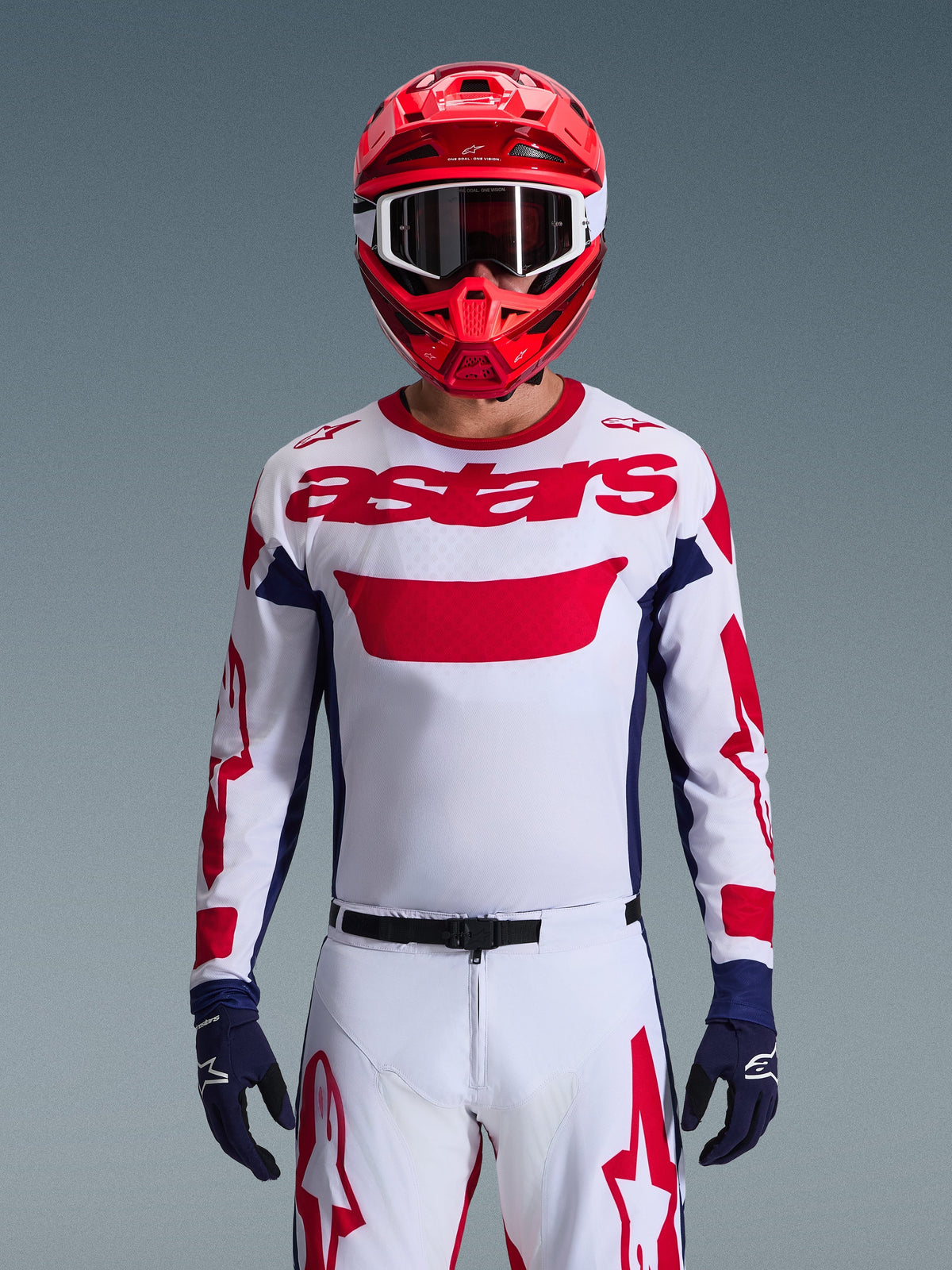 Piloto de motocross a usar a Camisola Alpinestars 2026 Racer Riway em Branco, Azul e Vermelho, com um grande logótipo 'astars' vermelho no peito, painéis laterais em azul marinho, calças de motocross brancas e vermelhas a condizer, capacete vermelho com óculos brancos e luvas azul marinho, conjunto completo de equipamento de corrida off-road