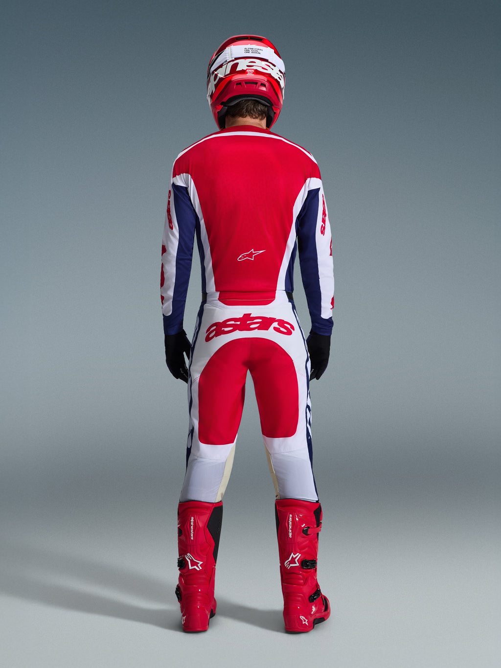 Piloto de motocross visto de costas a usar o conjunto Alpinestars Racer Riway 2026, camisola vermelha e azul com detalhes brancos, calças brancas com painéis vermelhos e logótipo 'astars' na zona do assento, coordenado com capacete vermelho, botas vermelhas e luvas pretas, equipamento de alta performance para todo-o-terreno