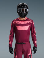 Piloto de motocross a usar o Conjunto de Equipamento Alpinestars 2026 Racer Riway, em grená e rosa, camisola com logótipo rosa em destaque no peito, calças a combinar com detalhes em preto e rosa, capacete integral preto com óculos e luvas pretas, fato de corrida off-road coordenado.