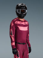Piloto de motocross a usar o Conjunto de Equipamento Alpinestars 2026 Racer Riway, camisola bordeaux e rosa com a marca 'astars' em destaque, calças a condizer com detalhes rosa, capacete e luvas pretas, fato profissional para motocross todo-o-terreno