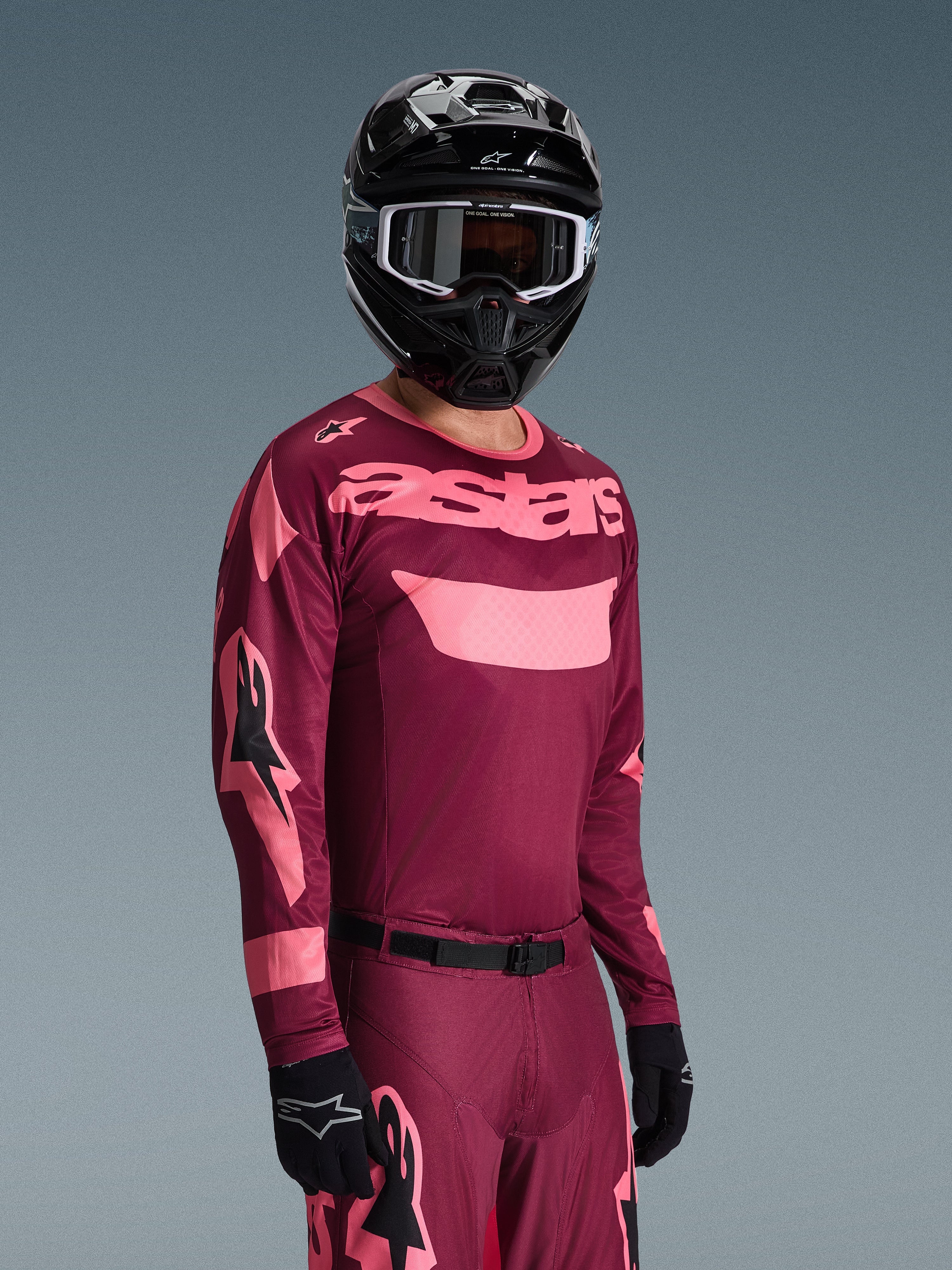 Piloto de motocross a usar o Conjunto de Equipamento Alpinestars 2026 Racer Riway, camisola bordeaux e rosa com a marca 'astars' em destaque, calças a condizer com detalhes rosa, capacete e luvas pretas, fato profissional para motocross todo-o-terreno