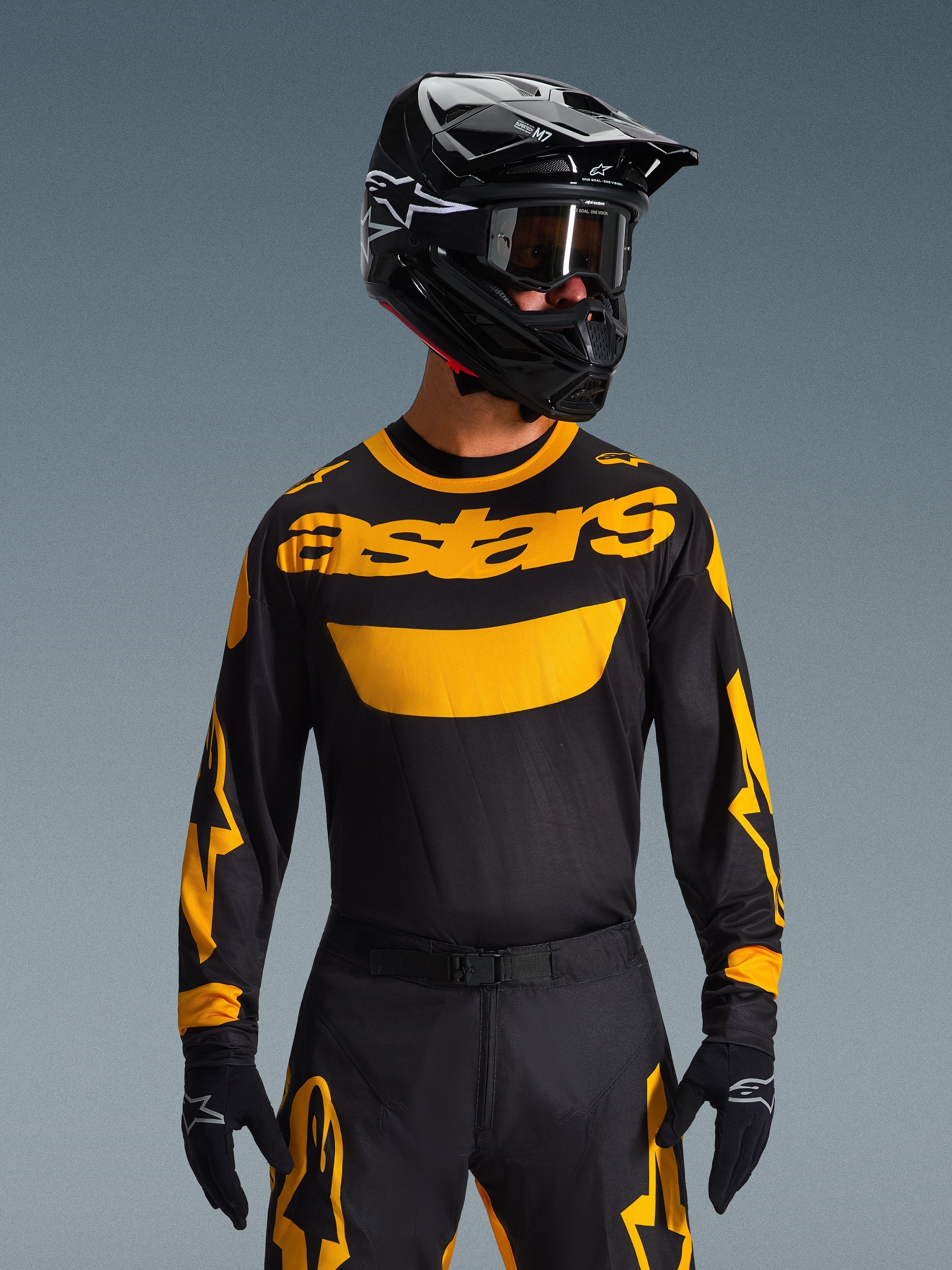 Piloto de motocross a usar a Camisola Alpinestars Racer Riway 2026 em amarelo e preto, com o logótipo "astars" amarelo em destaque no peito, calças coordenadas em preto e amarelo, capacete preto com óculos e luvas pretas, conjunto de equipamento profissional para todo-o-terreno