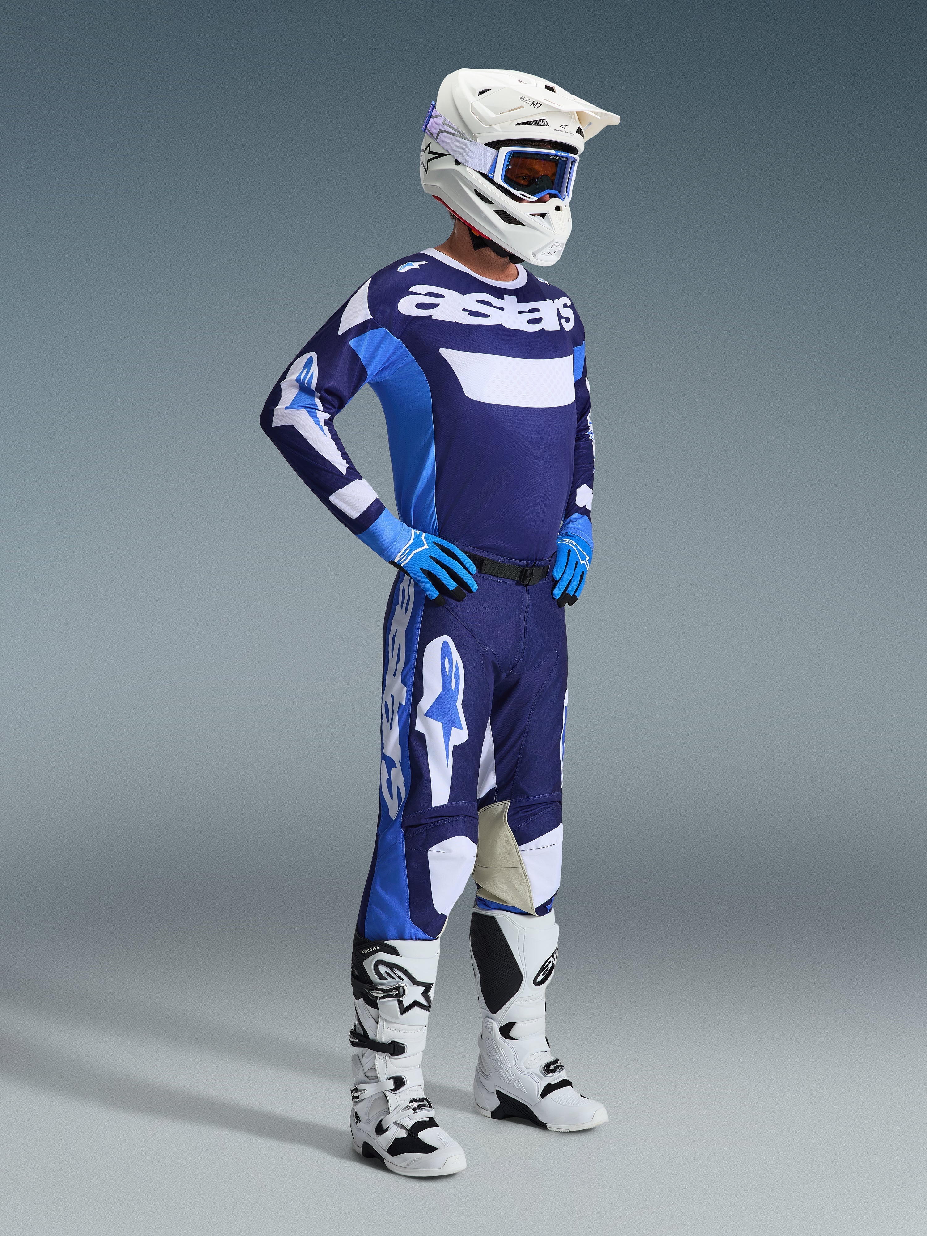 Piloto de motocross com o conjunto de equipamento Alpinestars 2026 Racer Riway em Azul UCLA e Branco, camisola azul e branca com a marca 'astars', calças a condizer, capacete branco com óculos azuis e botas de todo-o-terreno brancas, equipamento de corrida completo para motocross