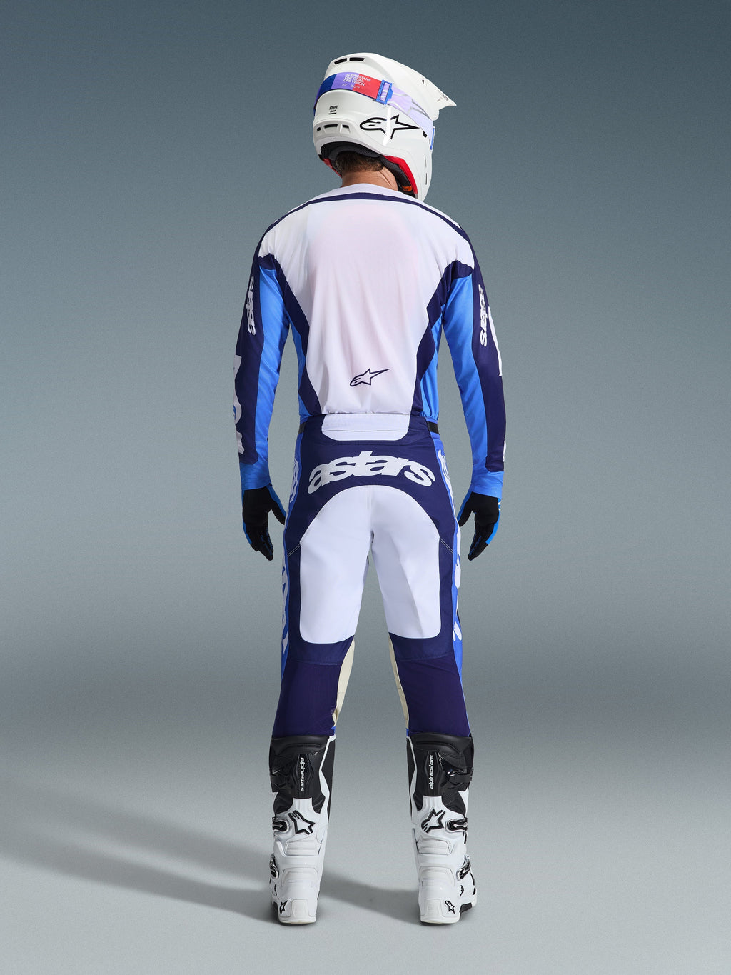 Vista traseira de um piloto de motocross a usar a Camisola e Calças Alpinestars 2026 Racer Riway em Azul Ucla e Branco, coordenado com um capacete branco, luvas pretas e botas de motocross brancas, equipamento de alta performance para todo-o-terreno