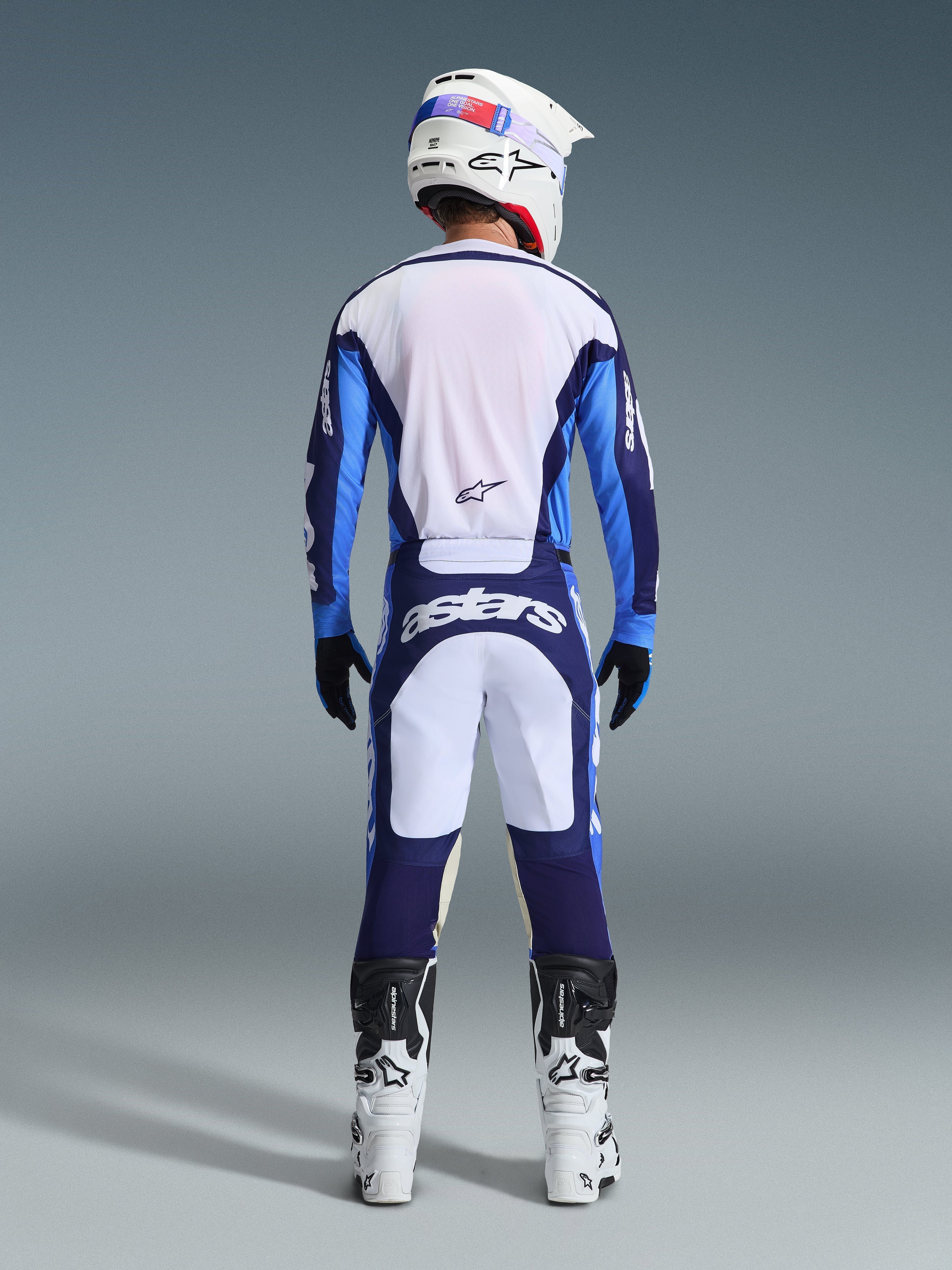 Vista traseira de um piloto de motocross a usar a Camisola e Calças Alpinestars 2026 Racer Riway em Azul Ucla e Branco, coordenado com um capacete branco, luvas pretas e botas de motocross brancas, equipamento de alta performance para todo-o-terreno