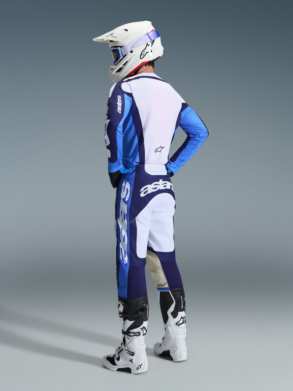 Piloto de motocross visto de costas e de lado a usar o conjunto de equipamento Alpinestars 2026 Racer Riway em azul UCLA e branco, com camisola azul e branca com a marca 'astars', calças a condizer, capacete de motocross branco com óculos azuis e botas brancas e pretas da série Tech