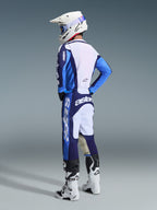Piloto de motocross visto de costas e de lado a usar o conjunto de equipamento Alpinestars 2026 Racer Riway em azul UCLA e branco, com camisola azul e branca com a marca 'astars', calças a condizer, capacete de motocross branco com óculos azuis e botas brancas e pretas da série Tech