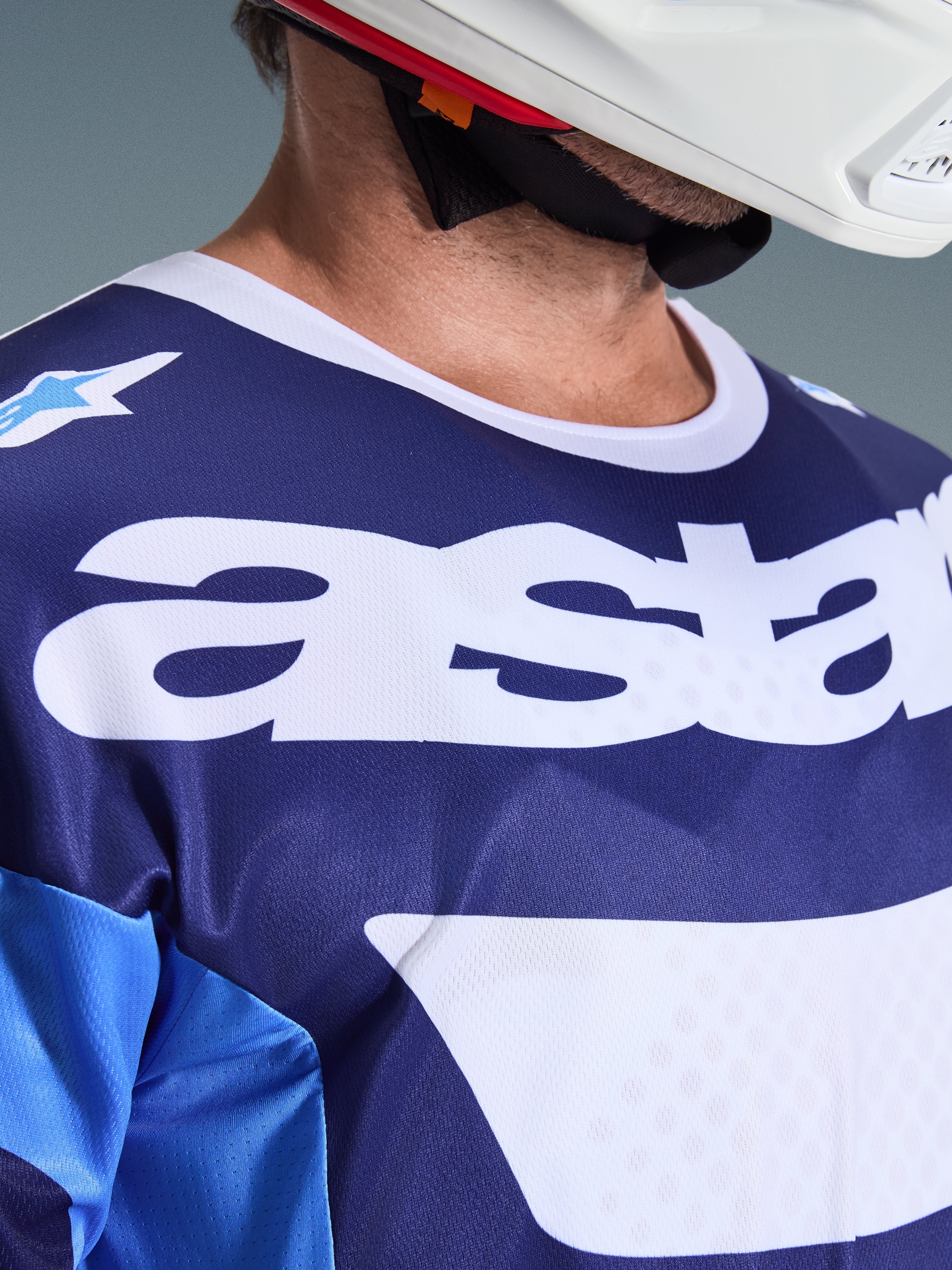 Alpinestars 2026 Racer Riway Jersey, camisola de motocross, azul UCLA e branco, vista de pormenor do peito com logótipo 'astars' de grandes dimensões em branco, gola branca e textura de malha que absorve a humidade, usada com um capacete branco