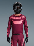 Piloto de motocross com a Camisola Alpinestars Racer Riway 2026 em Dark Red Coral, com corpo cor de vinho e logótipo 'astars' no peito em rosa coral, combinada com calças de motocross a condizer, capacete e luvas pretas, conjunto de equipamento profissional para todo-o-terreno.