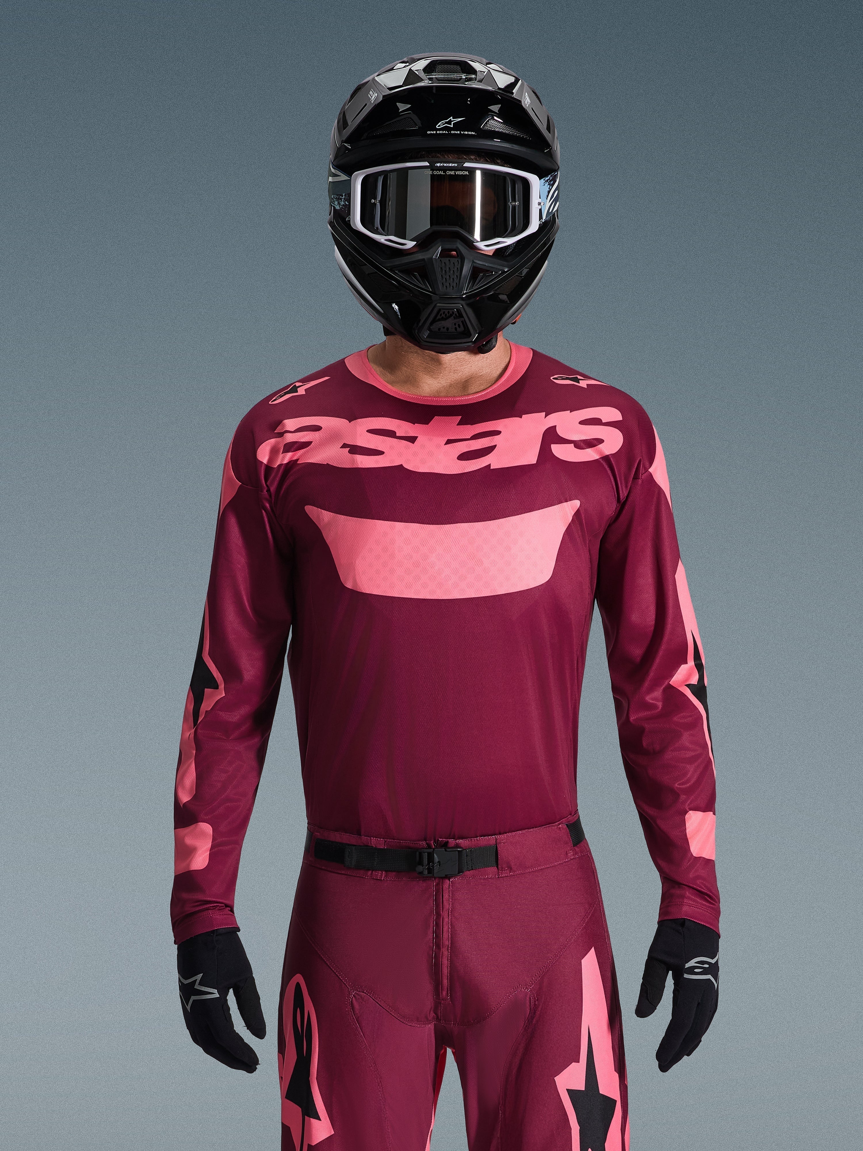 Piloto de motocross com a Camisola Alpinestars Racer Riway 2026 em Dark Red Coral, com corpo cor de vinho e logótipo 'astars' no peito em rosa coral, combinada com calças de motocross a condizer, capacete e luvas pretas, conjunto de equipamento profissional para todo-o-terreno.