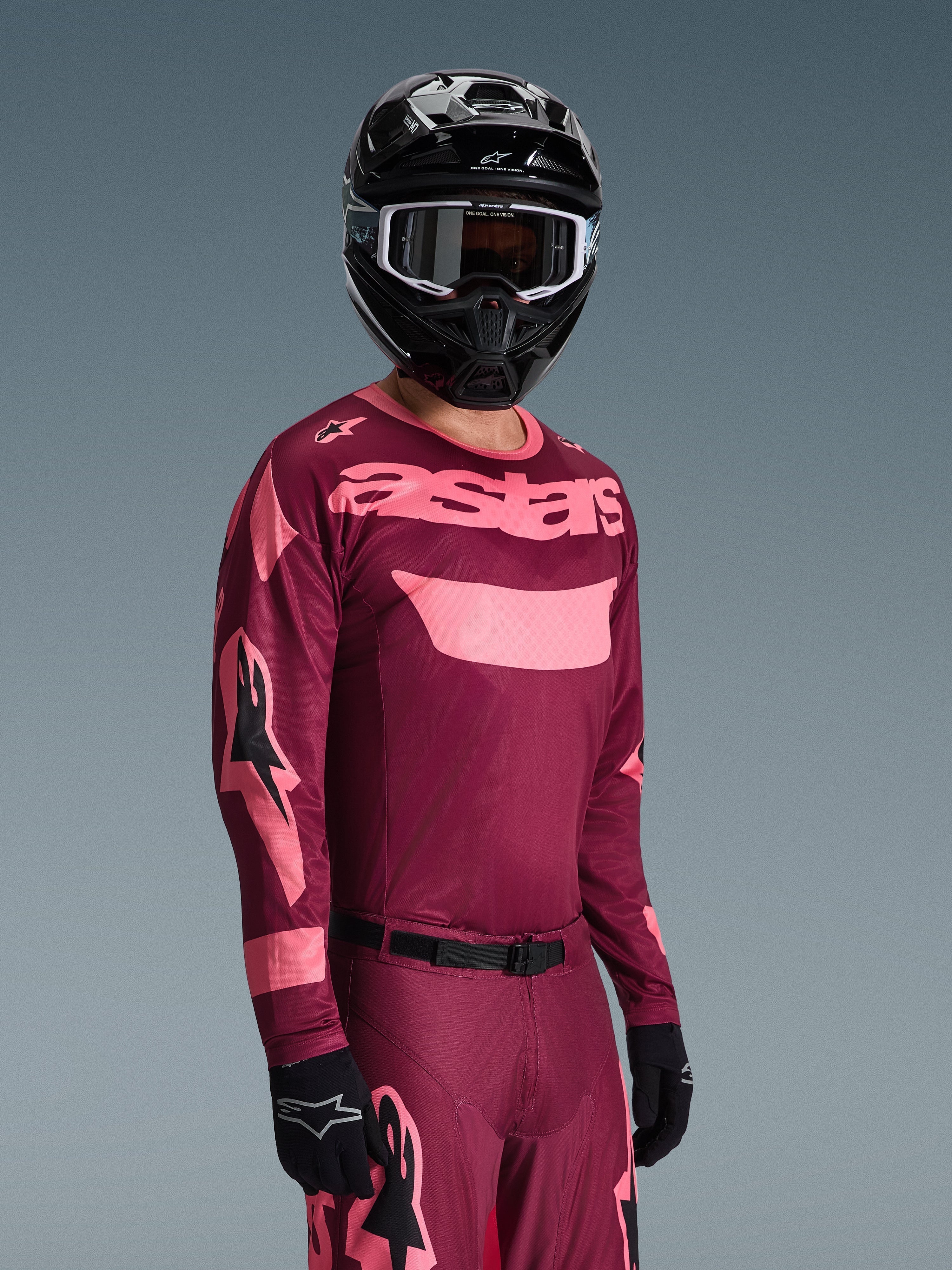 Piloto de motocross a usar a camisola Alpinestars 2026 Racer Riway em Dark Red Coral com calças a condizer, com a marca 'astars' em cor-de-rosa coral no peito, acompanhado de capacete e luvas pretas, conjunto de equipamento de corrida todo-o-terreno.