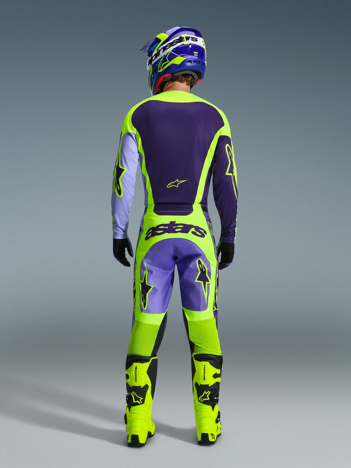 Piloto de motocross com o conjunto de equipamento Alpinestars Racer Portl 2026, vista traseira, camisola roxo escuro e amarelo néon com mangas lavanda, calças a condizer com marca 'astars', botas amarelo néon vibrante, equipamento profissional para todo-o-terreno