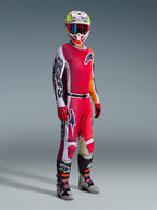 Piloto de motocross com equipamento de corrida completo, camisola Alpinestars Racer Portl 2026 em vermelho e cinzento claro, calças de motocross a condizer com detalhes em branco e verde escuro, capacete multicolorido com óculos vermelhos, botas cinzentas e pretas com detalhes laranja e roxo, luvas vermelhas, conjunto coordenado para corridas todo-o-terreno