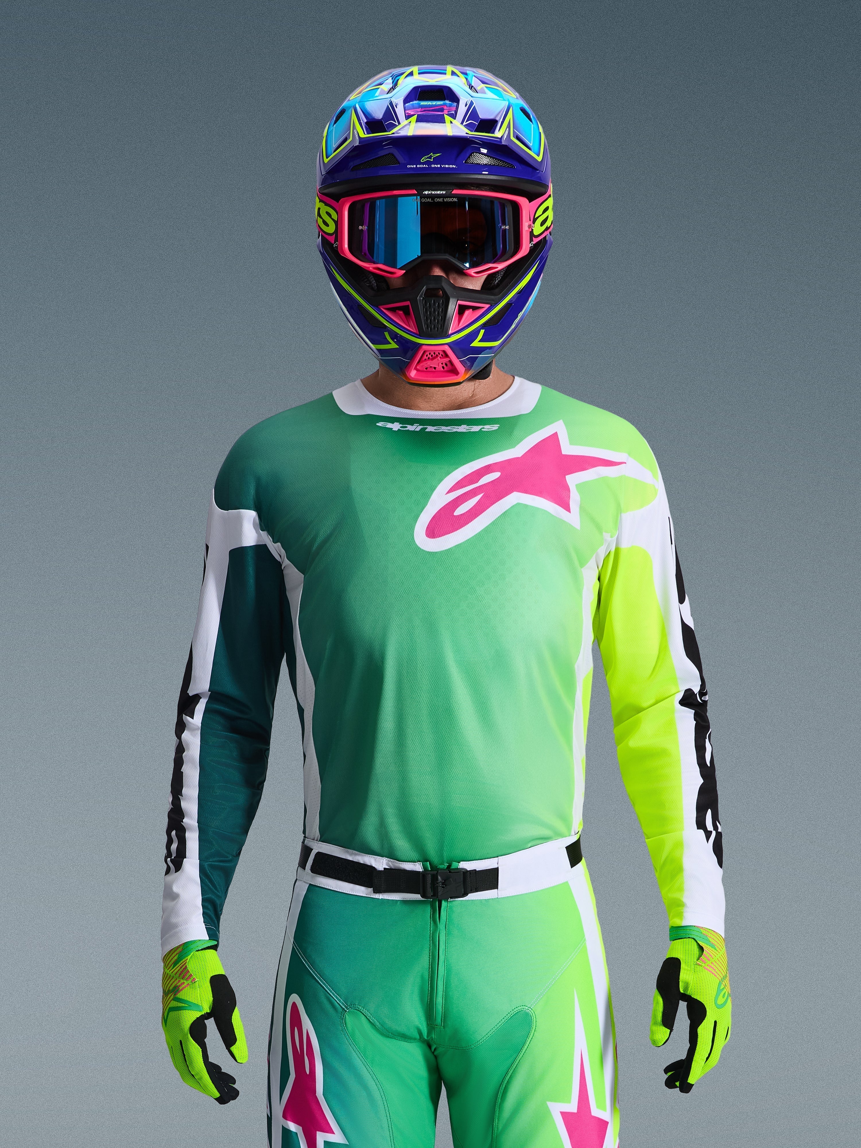 Alpinestars 2026 Racer Portl Jersey, camisola de motocross, verde, branco e preto com um grande logótipo Alpinestars rosa no peito, usada por um piloto com um capacete multicolorido vibrante e óculos a condizer, vestuário profissional de competição off-road