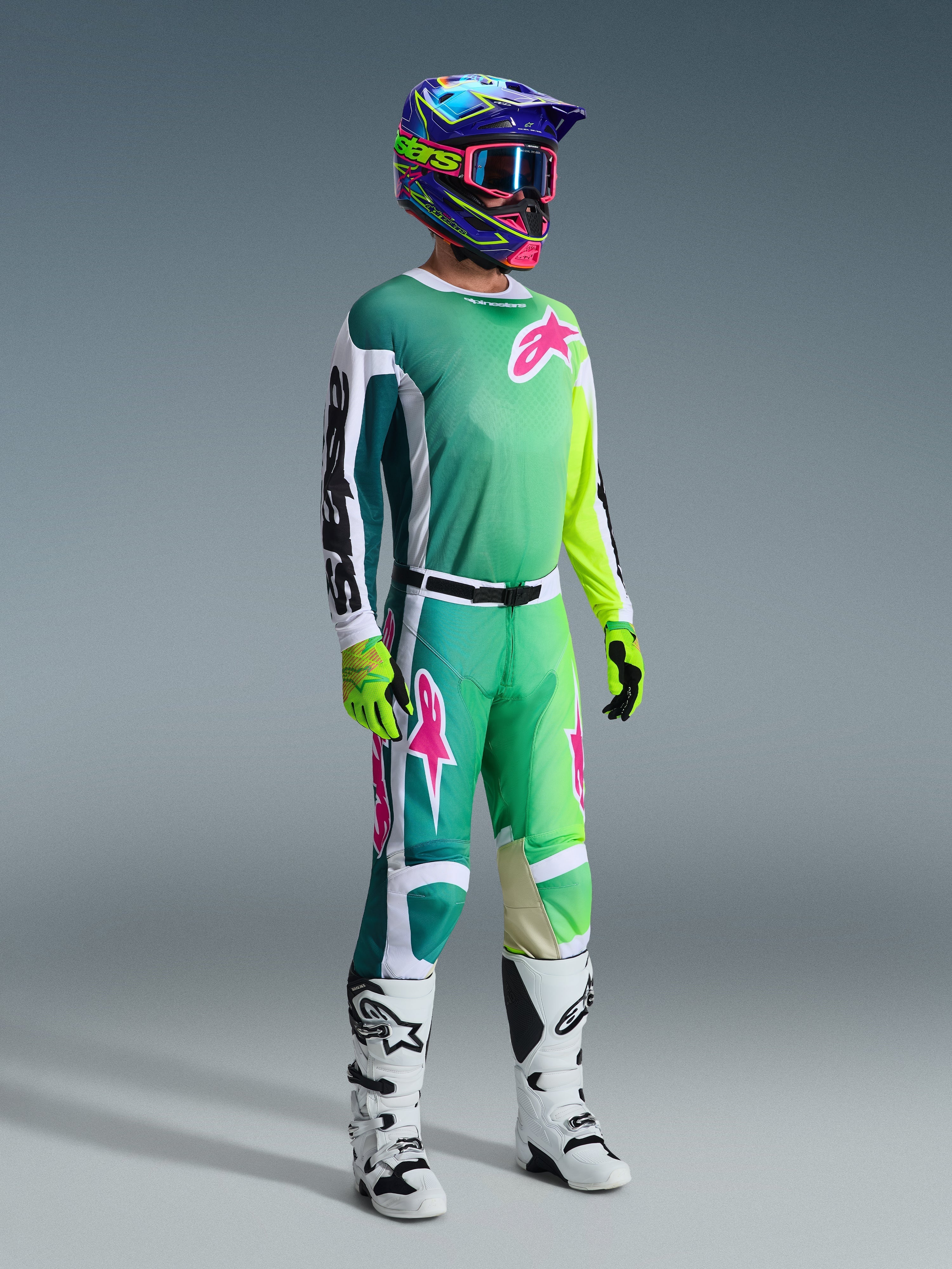 Piloto de motocross com equipamento de competição completo, Camisola Racer Portl 2026 em verde com mangas brancas e pretas e logótipo cor-de-rosa, calças verdes a condizer com detalhes cor-de-rosa, capacete multicolorido com óculos, botas brancas e pretas e luvas verdes, conjunto coordenado para corridas off-road