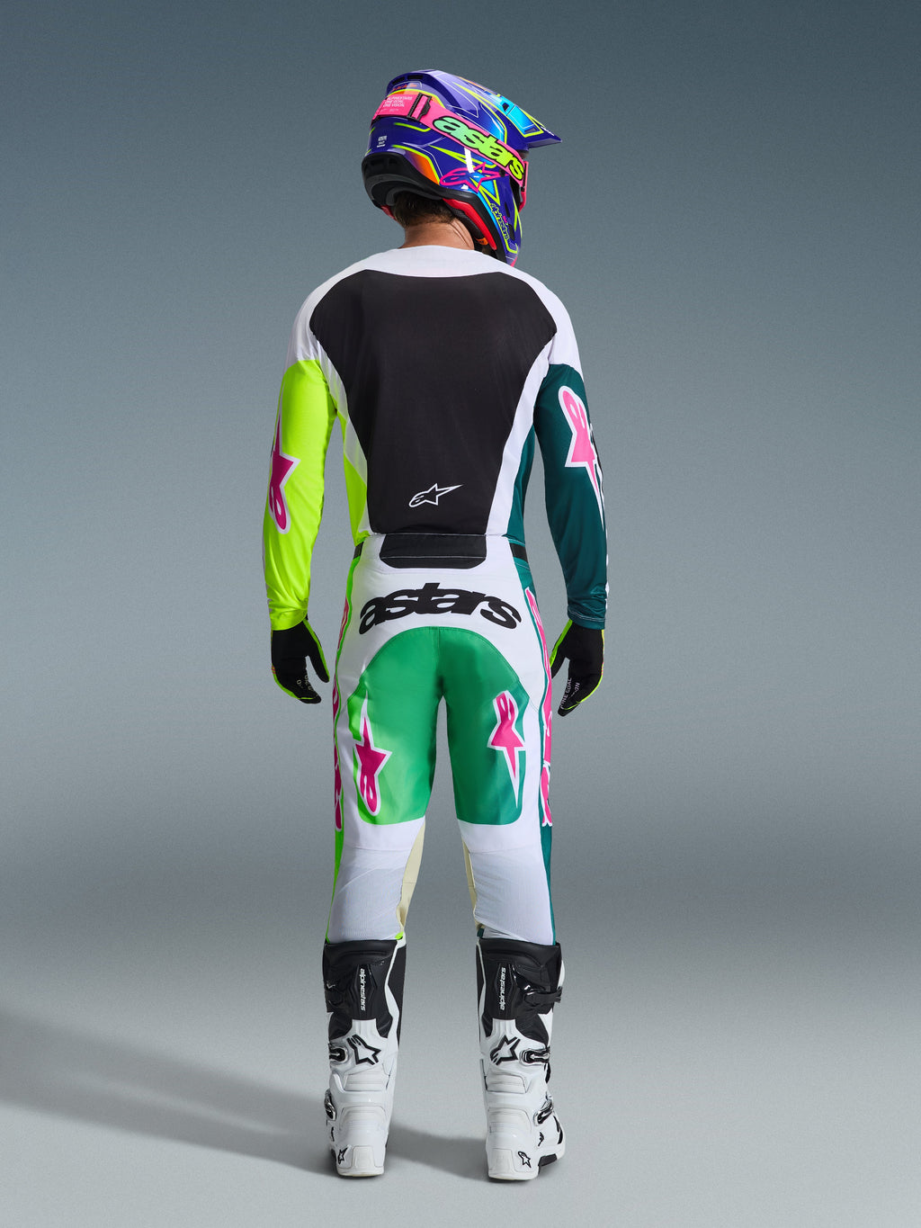 Vista traseira de um piloto de motocross a usar o conjunto de equipamento Alpinestars 2026 Racer Portl, camisola branca com painel traseiro preto e mangas em amarelo néon e verde escuro com estrelas cor-de-rosa, calças brancas com detalhes em verde e verde-azulado e logótipos de estrelas cor-de-rosa, acompanhado por capacete colorido, luvas pretas e botas brancas e pretas.