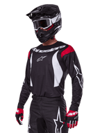 Motocross rider wearing Alpinestars 2025 Fluid Haul Gear Set, black jersey with red shoulder panels and white graphics, matching black pants with white star logos, red and black gloves, black helmet with white accents and goggles, coordinated motocross apparel.